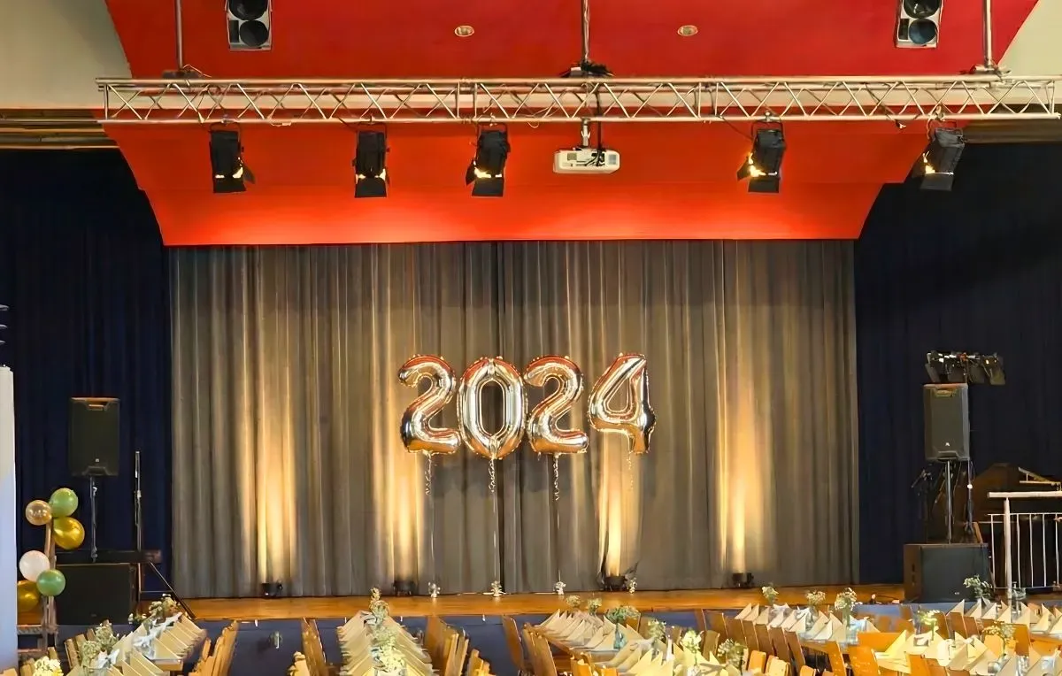 Festlich gedeckte Tische vor einer Bühne mit Lautsprechern, Traversen und warmer Beleuchtung beim Abiball